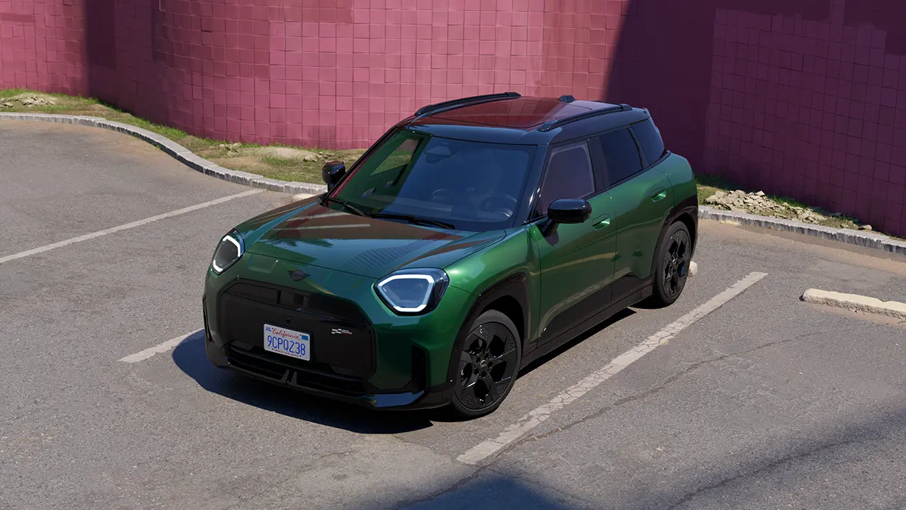 Frontansicht eines vollelektrischen MINI Aceman in British Racing Green, der auf einem Parkplatz parkiert, mit einer rosafarbenen Wand im Hintergrund
