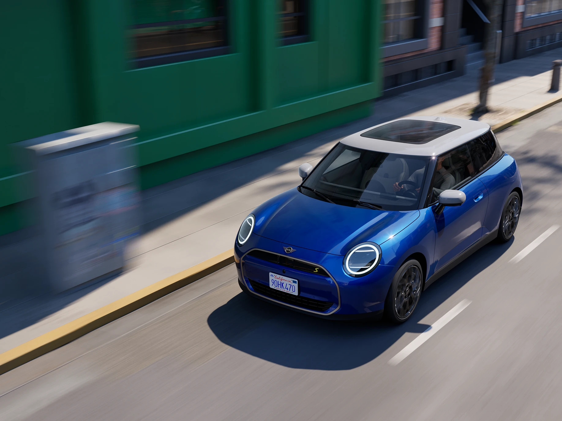 Draufsicht auf den vollelektrischen MINI Cooper in Blazing Blue mit weißem Dach beim Fahren auf einer Straße. 