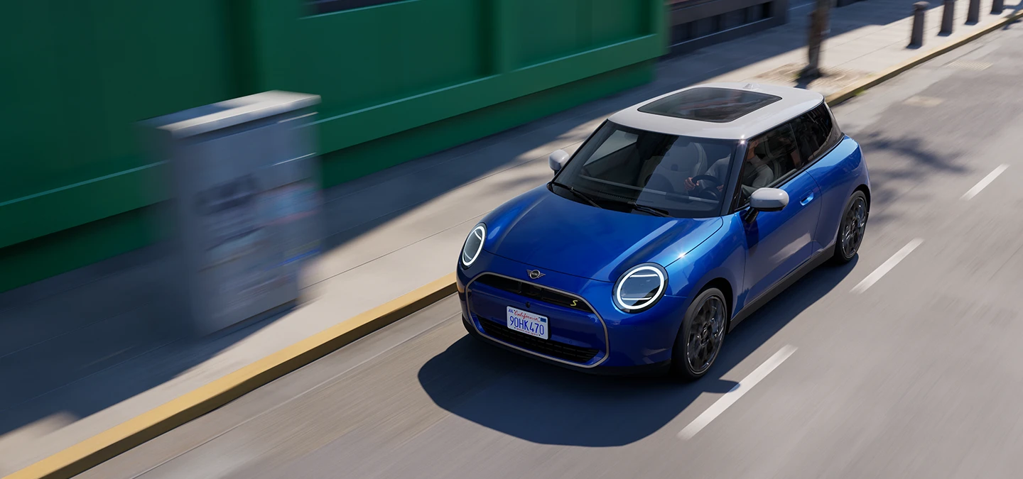 Draufsicht auf den vollelektrischen MINI Cooper in Blazing Blue mit weißem Dach beim Fahren auf einer Straße. 