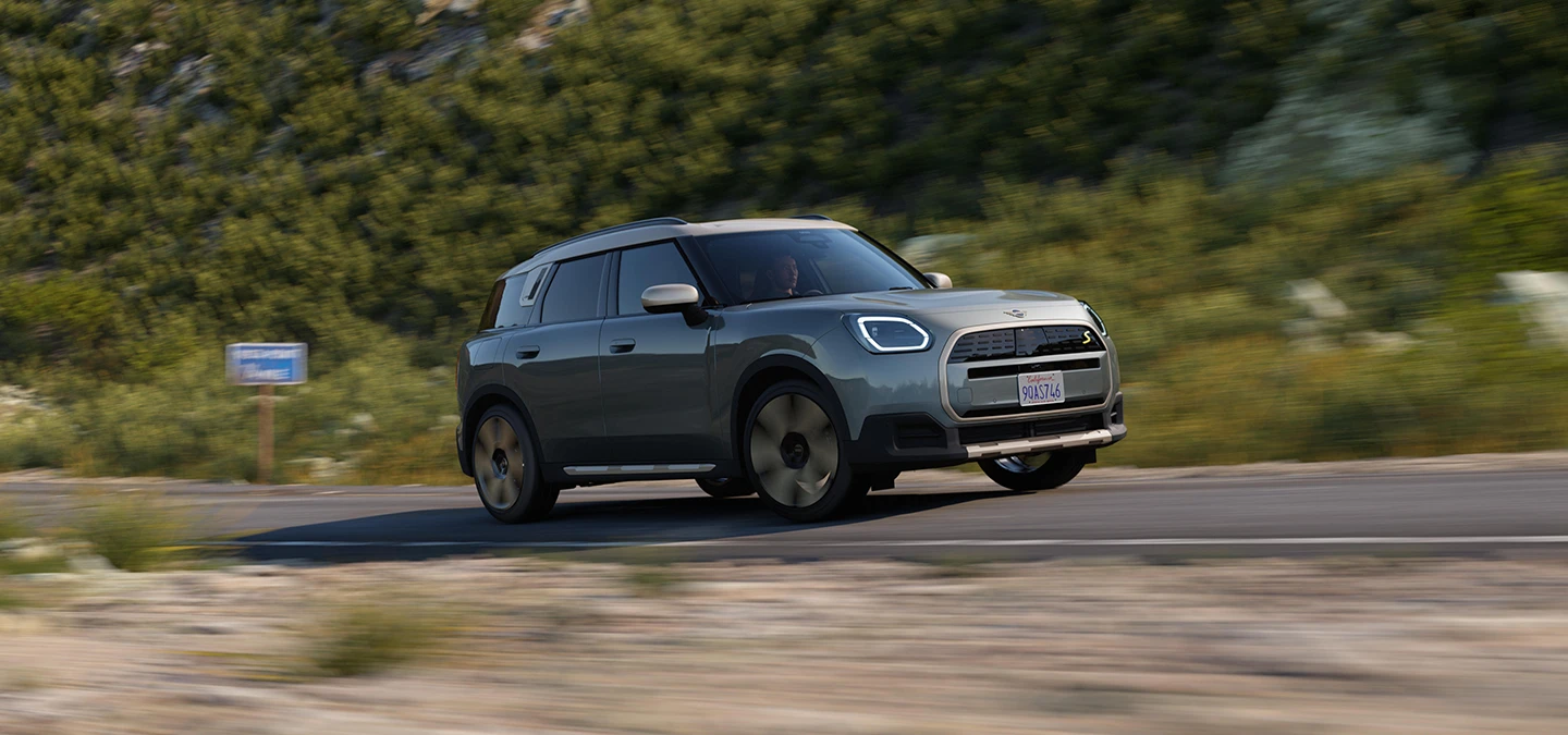 Seitenansicht eines im Gelände fahrenden vollelektrischen MINI Countryman in Smokey Green mit Dach in Vibrant Silver.