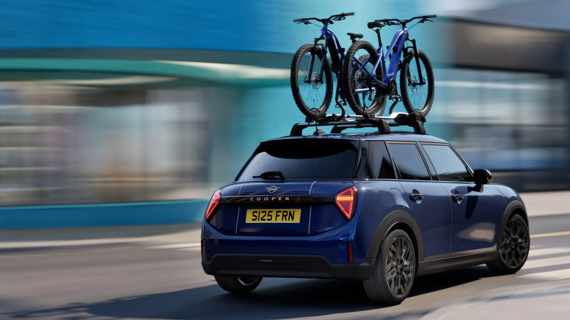 Dreiviertelansicht eines MINI Aceman in Indigo Sunset Blue von hinten, ausgestattet mit einem MINI Fahrraddachträger, auf dem zwei Mountainbikes befestigt sind. 